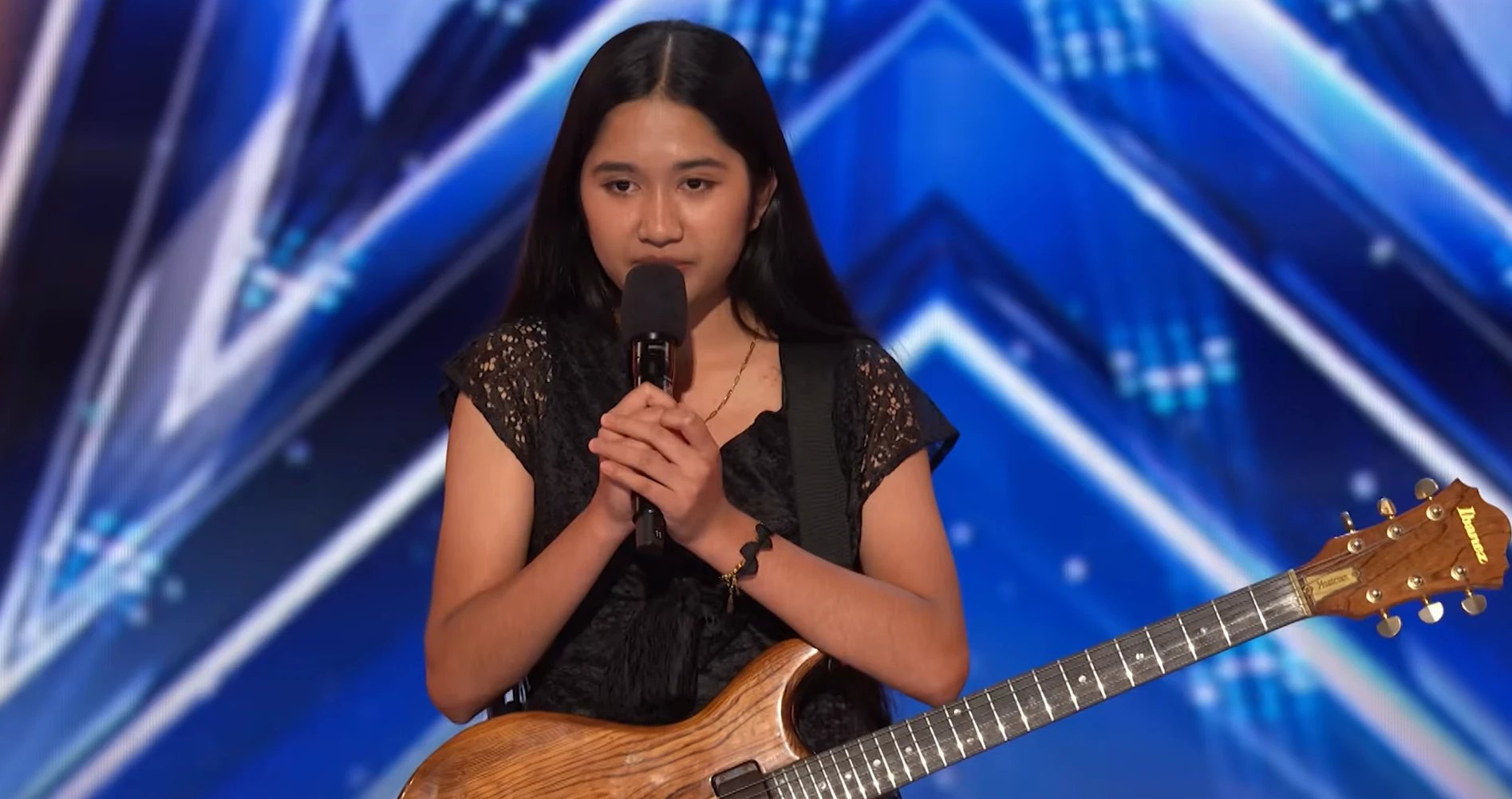 Nene Royal standing on the America's Got Talent stage holding the microphone, vintage Ibanez guitar across her body, blue AGT backdrop behind her