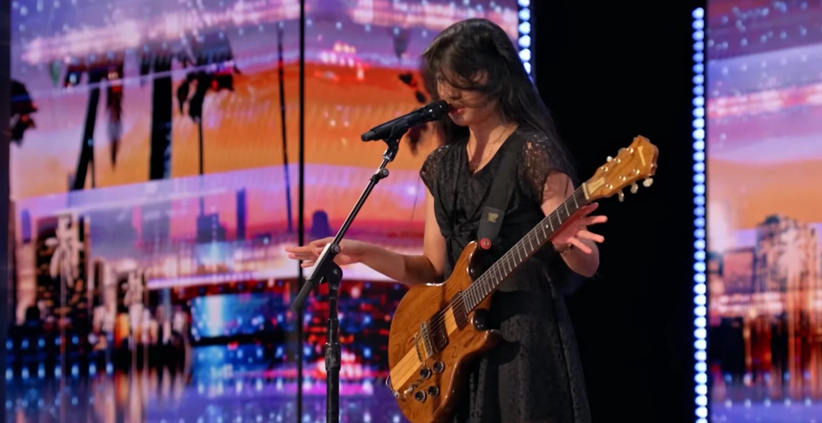 Nene Royal playing her Ibanez guitar at the microphone stand on the AGT stage, city skyline backdrop behind her, full-body performance shot