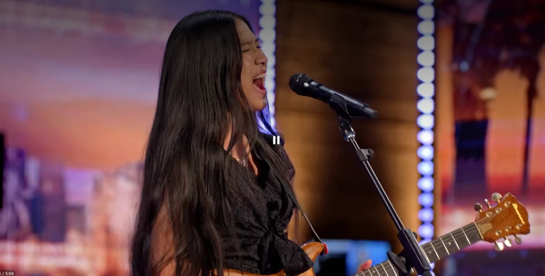 Close-up side profile of Nene Royal singing with intense passion at the AGT microphone, mouth open, eyes closed, guitar headstock visible, warm stage lighting